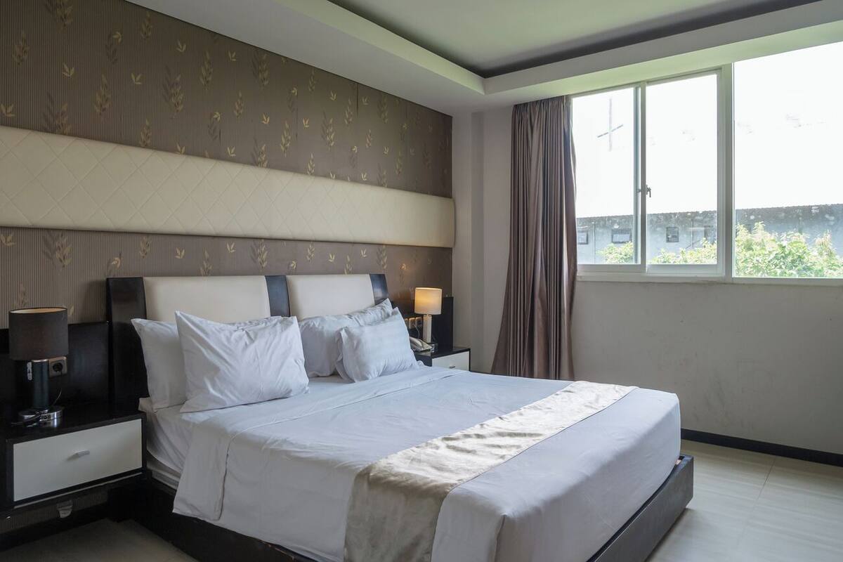 Una habitación de hotel con una cama grande, una mesita de noche con lámpara y una ventana con cortinas.