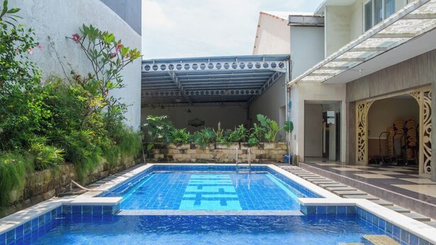 A swimming pool with a tiled deck, surrounded by greenery and a covered area with a patterned roof. - Princess Keisha By SECOM (Denpasar)