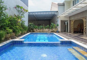 A swimming pool with a tiled deck, surrounded by greenery and a covered area with a patterned roof. - Princess Keisha By SECOM (Denpasar)