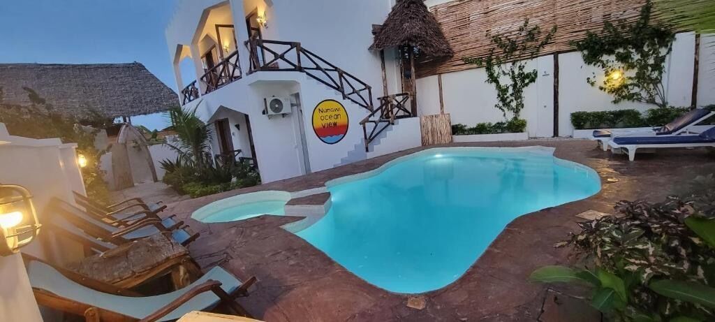 A swimming pool area with lounge chairs, a thatched-roof structure, and a sign reading "Nuwiwi ocean view."