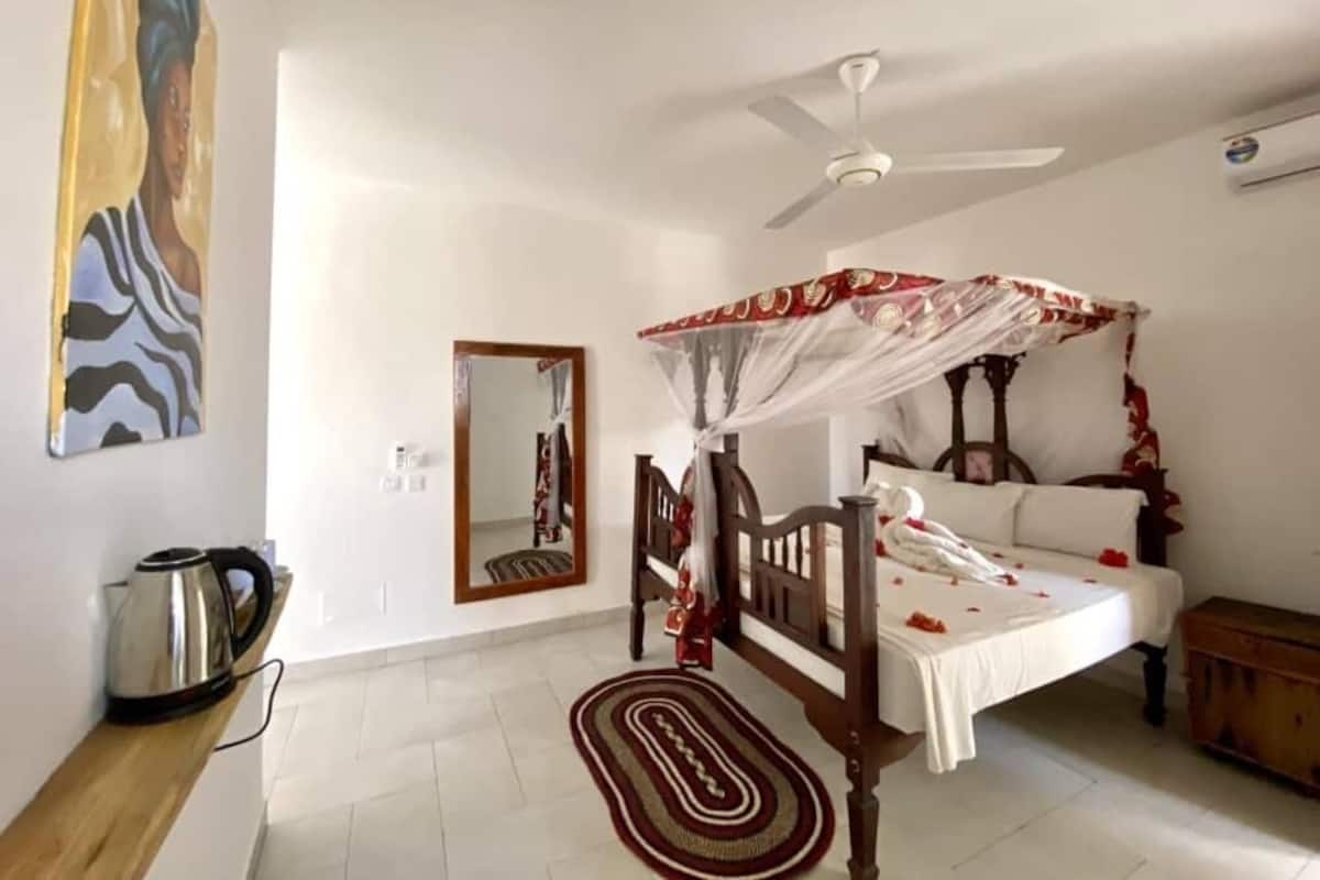 Un dormitorio con cama con dosel, ventilador de techo, mesita de noche con tetera y espejo en la pared.