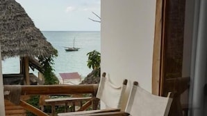 Une terrasse en bois offrant une vue sur la mer, un parasol en chaume et une chaise en osier avec un coussin.