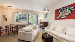A modern living room with a white sofa, a coffee table, a dining table with chairs, and a large window.