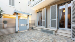 A residential building with a tiled patio, a closed garage door, and a set of steps leading to a door with shutters.