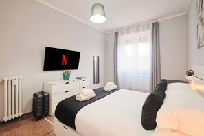 A modern hotel room with a large bed, a flat-screen TV mounted on the wall, a bedside table with a vase, and a window with curtains.