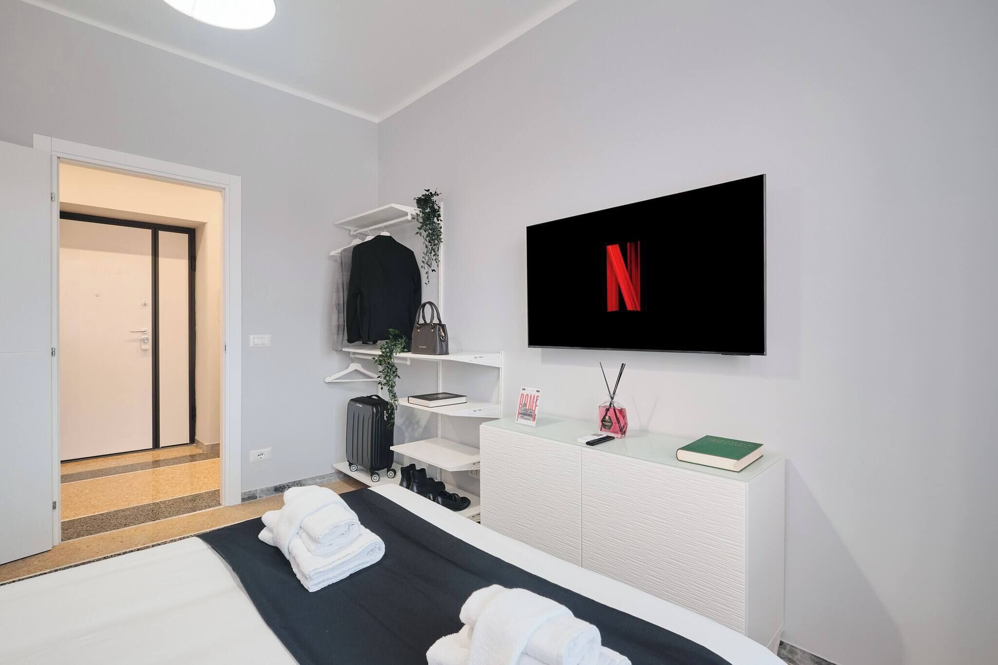 A modern hotel room with a bed, a TV mounted on the wall, a wardrobe, a desk with a plant, and a suitcase.