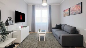 Un salon moderne avec un canapé gris, une table basse blanche, un téléviseur fixé au mur et une grande fenêtre avec des rideaux.