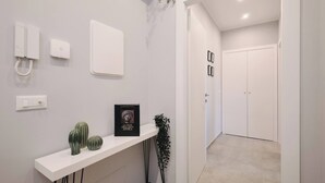 Un couloir moderne avec une console blanche, un tableau encadré et un cactus. On y trouve des interrupteurs fixés au mur et un téléphone. Une porte donne accès à une autre pièce.