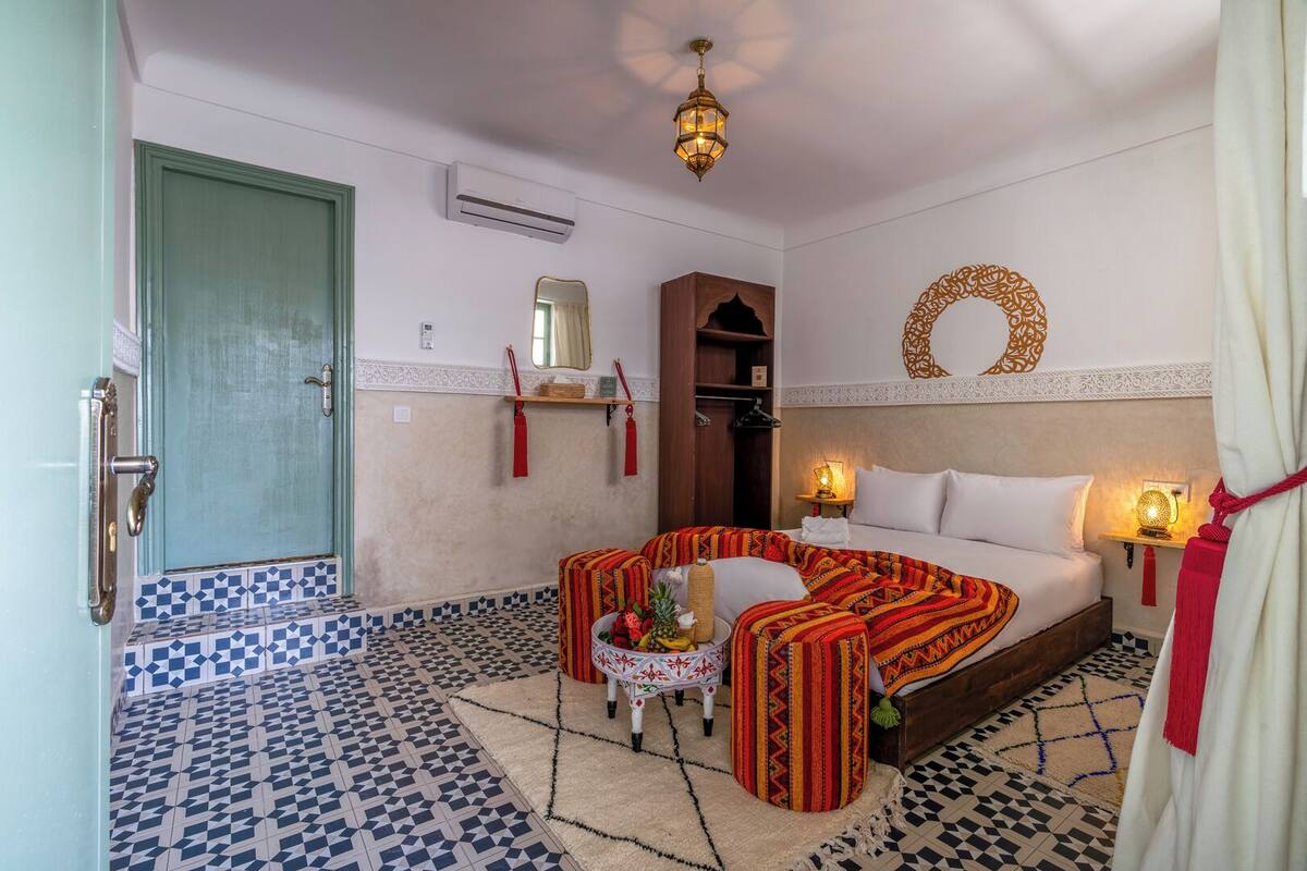 A hotel room with a bed, a small table with a lamp, a decorative mirror, and a wall-mounted air conditioner.