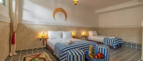 A hotel room with two beds, a decorative wall hanging, a patterned rug, and a fruit bowl on the floor.
