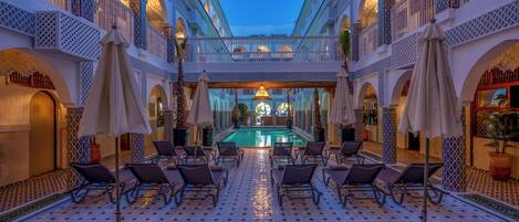 A luxurious indoor pool area with multiple lounge chairs and umbrellas, surrounded by ornate architecture and a central pool.