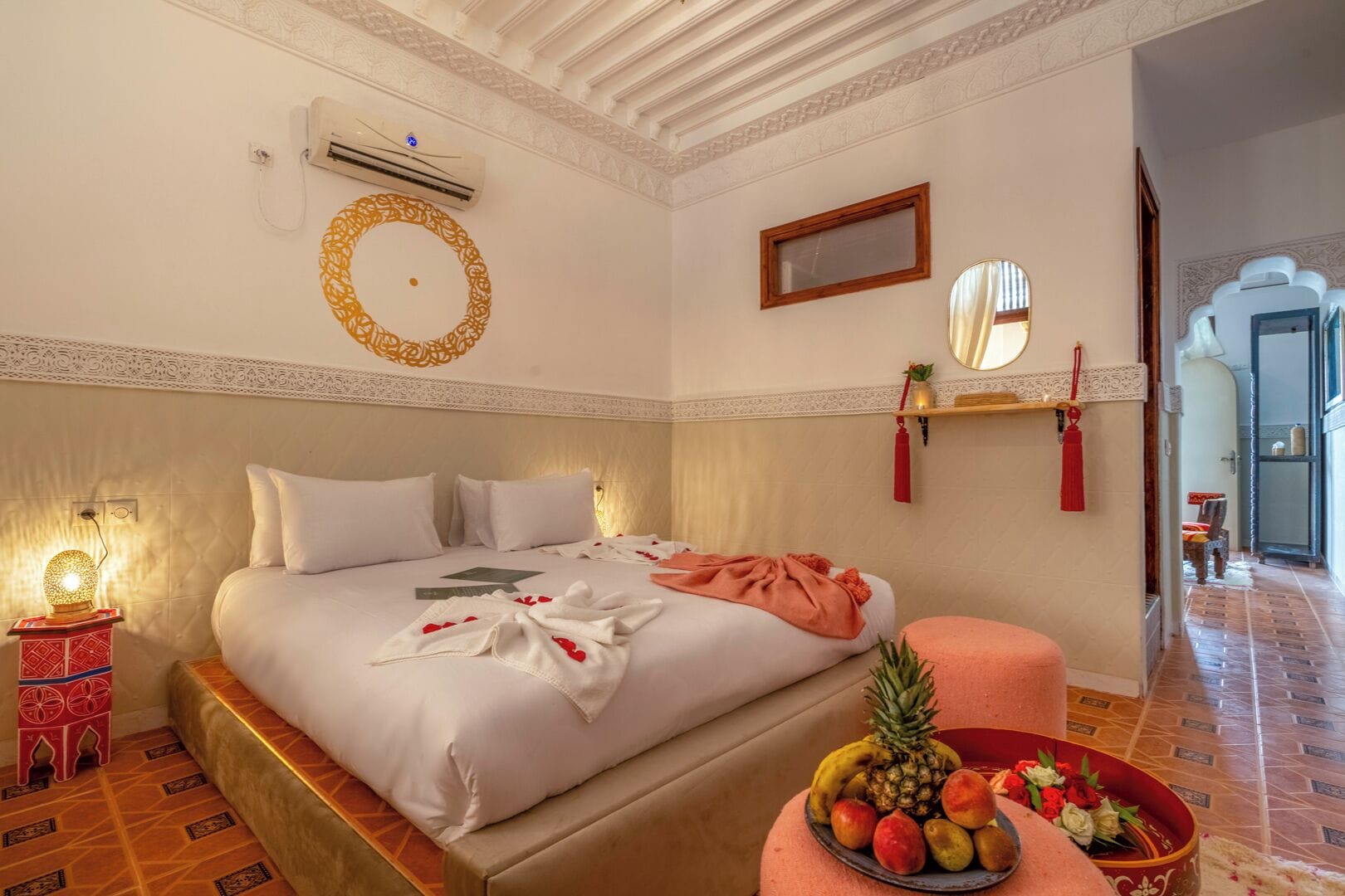 A hotel room with a large bed, a bedside table with a lamp, a wall-mounted air conditioner, a decorative mirror, and a fruit bowl on the floor.