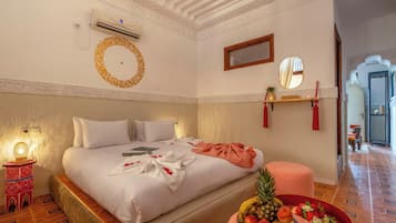 A hotel room with a large bed, a bedside table with a lamp, a wall-mounted air conditioner, a decorative mirror, and a fruit bowl on the floor.