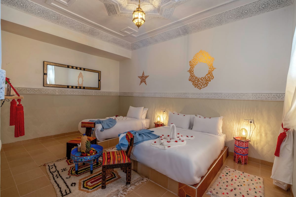 A hotel room with two beds, a mirror, decorative wall art, a small table with a fruit bowl, and a chair.