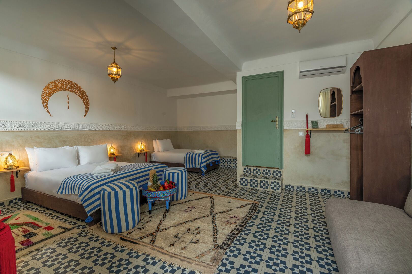 Habitación de hotel con dos camas, un sofá, una mesita con fruta, alfombras estampadas y apliques decorativos en la pared.