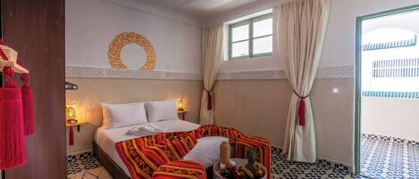A hotel room with a bed, a small table with fruit, a decorative wall piece, and a view of the outside.