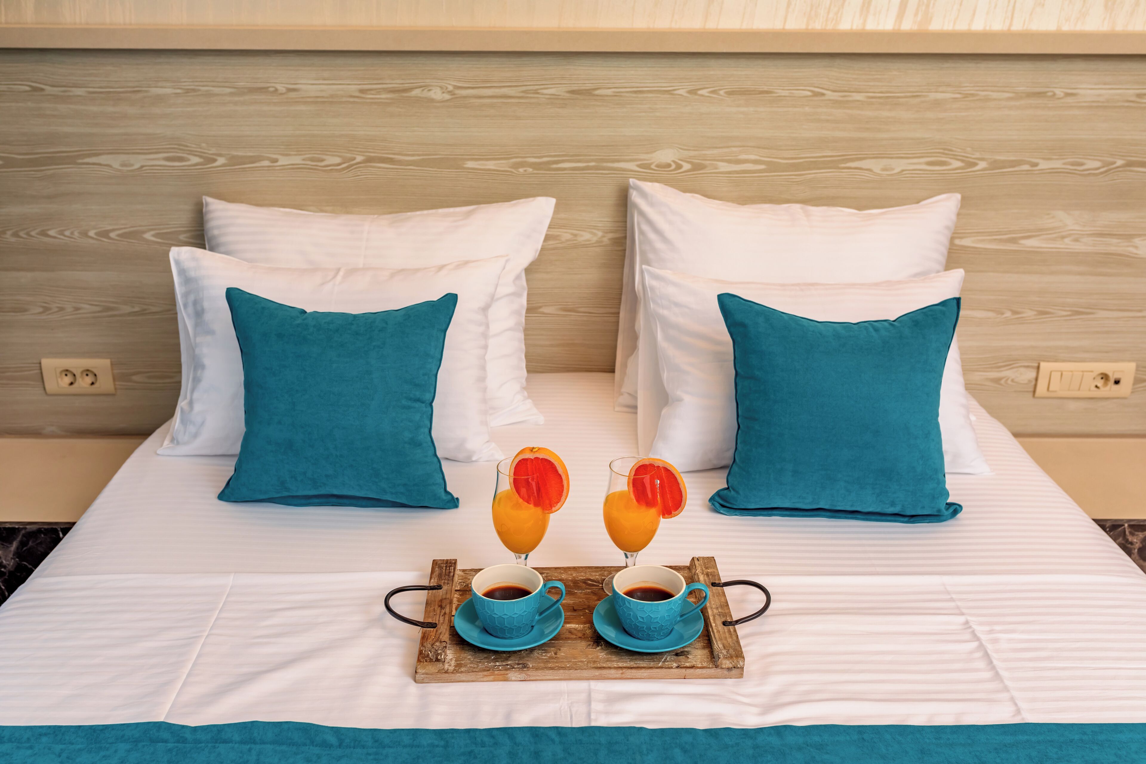 Una cama bien hecha con almohadas blancas y color turquesa, una bandeja con dos tazas de café y rodajas de naranja, y un cabecero de madera.