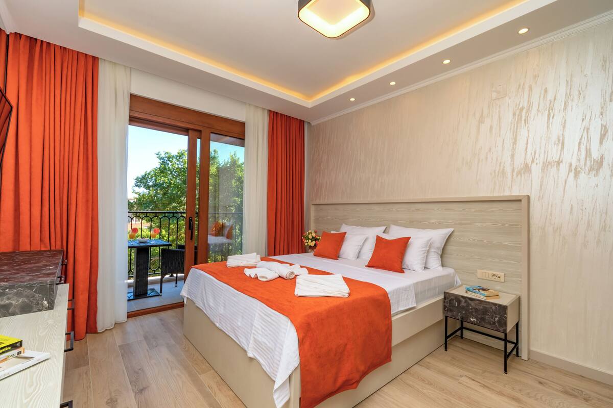 A modern hotel room with a large bed, orange curtains, a balcony with a table and chairs, and a view of greenery outside.