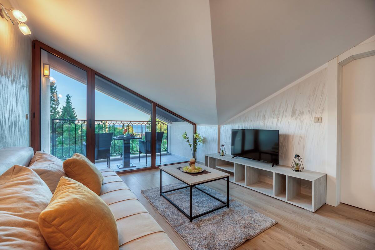 A modern living room with a large sofa, a coffee table, a TV, and a balcony with outdoor furniture.