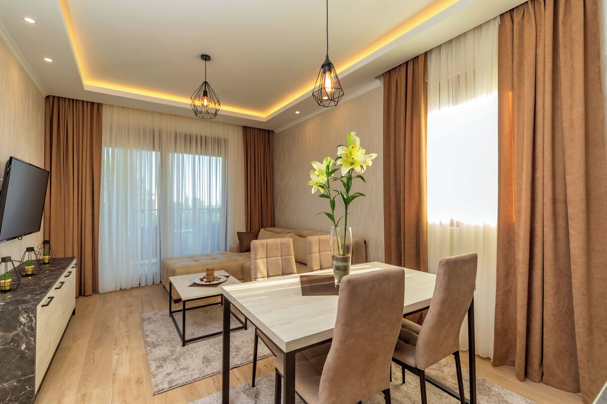 A modern living room with a dining area, featuring a flat-screen TV, a sofa, a coffee table, and a vase with flowers.
