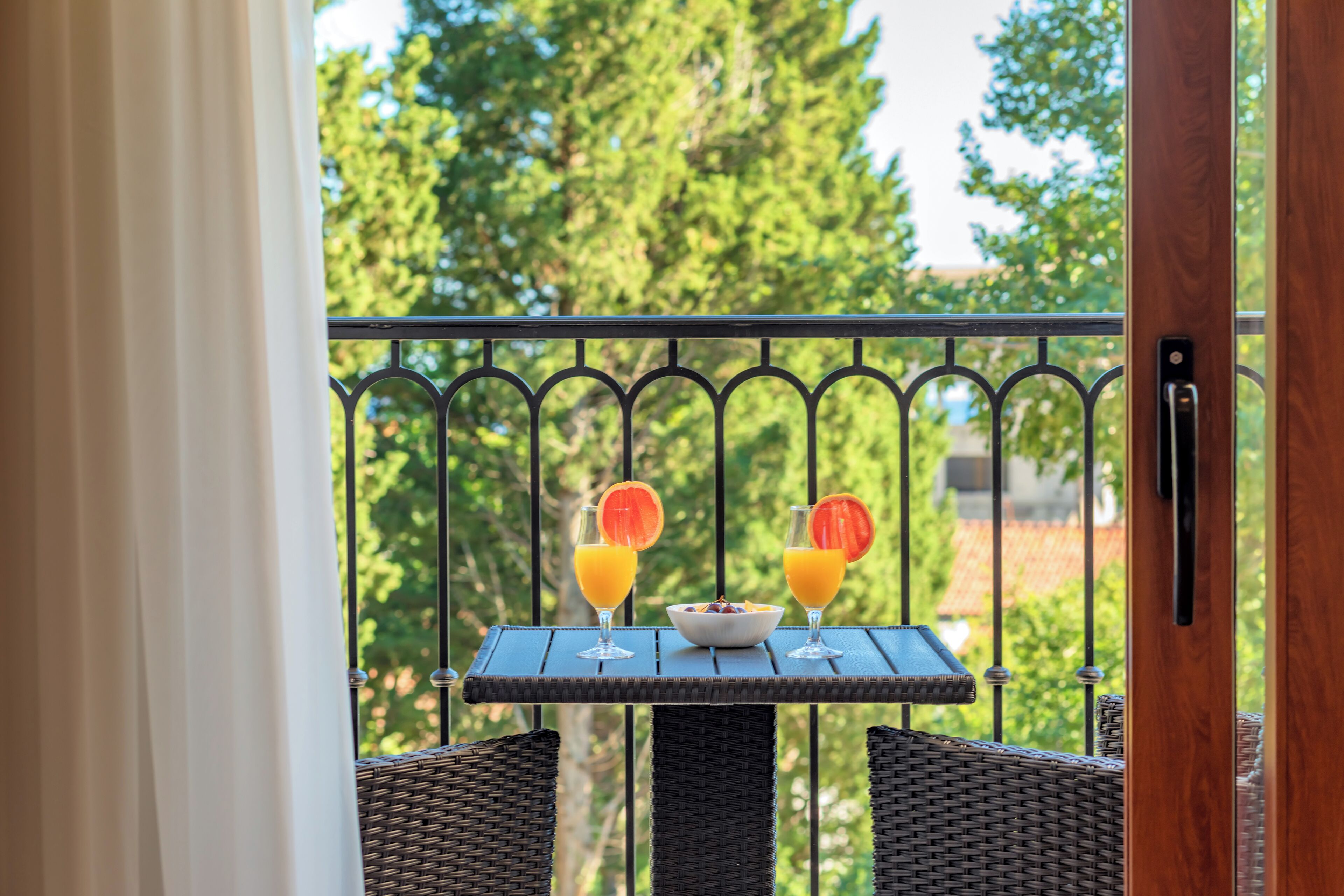 Un balcón con sillones de mimbre, una mesa con dos vasos de jugo de naranja y un bol de fruta, y vistas a árboles.