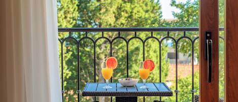 Un balcón con sillones de mimbre, una mesa con dos vasos de jugo de naranja y un bol de fruta, y vistas a árboles.