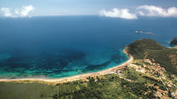 Vista aérea de un pueblo costero con playa, aguas azules cristalinas y un paisaje montañoso.