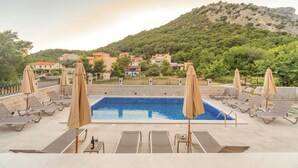 Zona de piscina con sillones de descanso y sombrillas, rodeada de edificios y una colina.