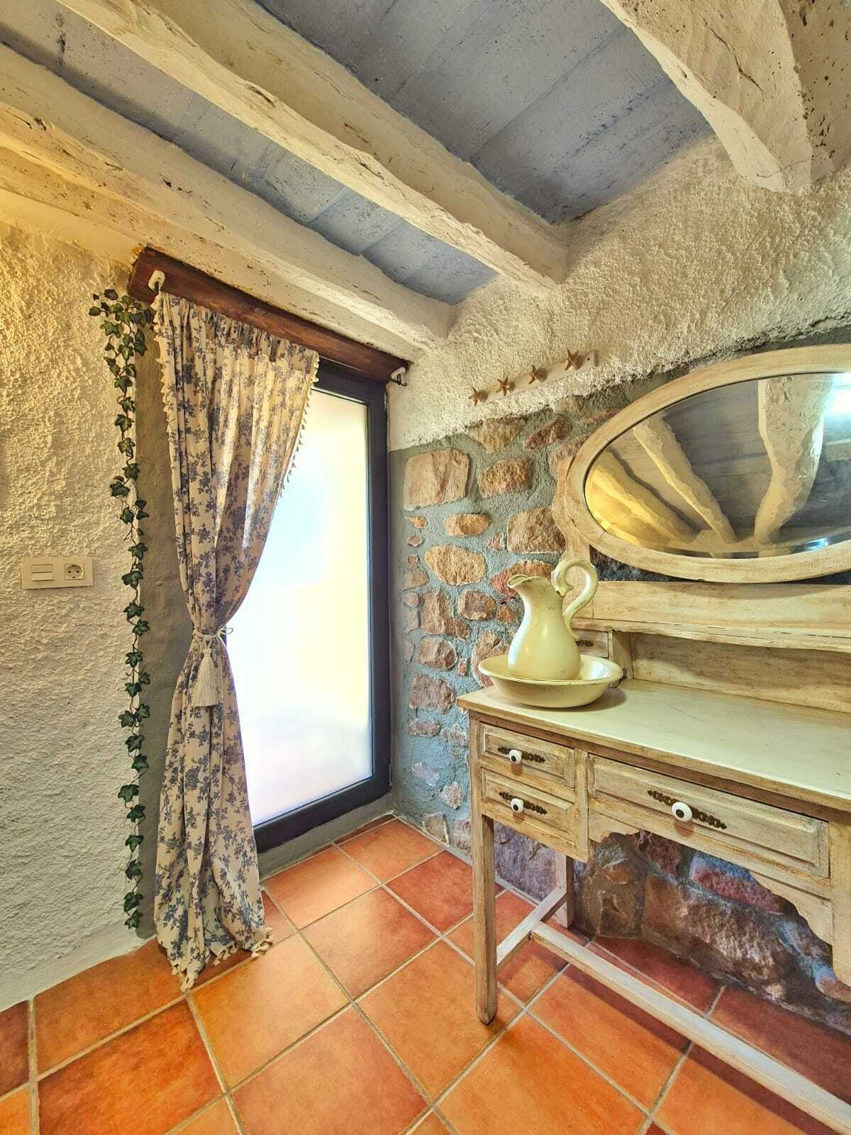 A rustic bathroom with a stone wall, a wooden vanity with a mirror, and a window with a curtain.