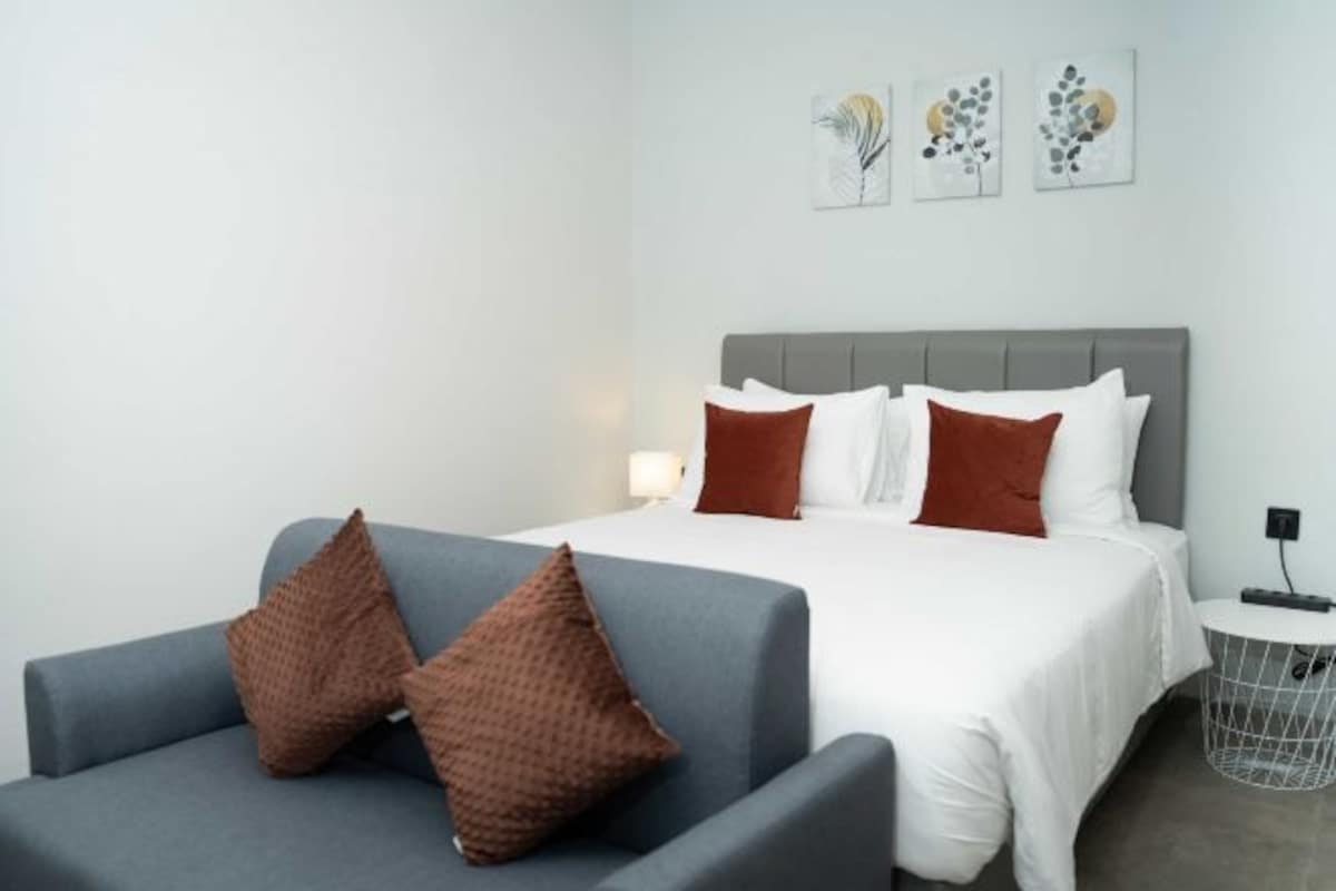 Une chambre d’hôtel moderne avec un canapé gris, un lit avec des oreillers blancs et bruns, et une petite table d’appoint avec une lampe.