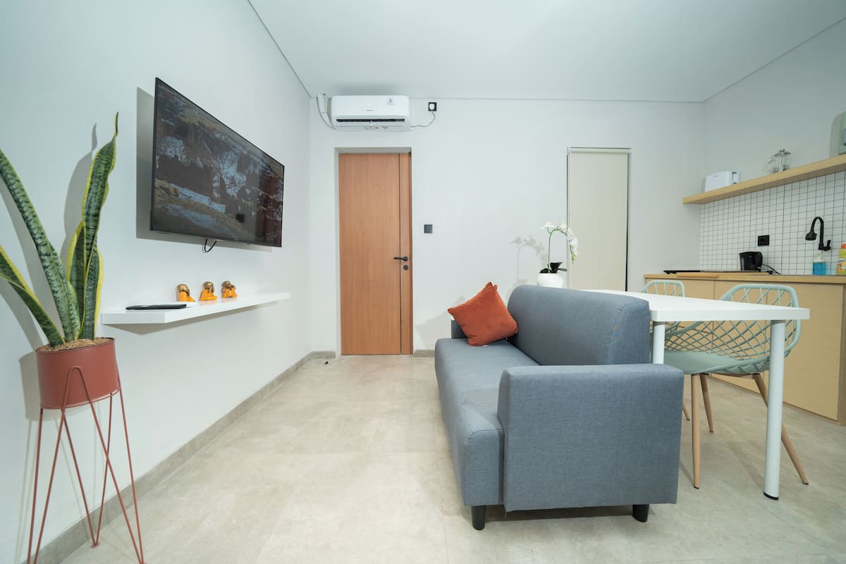 Een moderne woonkamer met een grijze bank, een flatscreen televisie, een potplant en een klein schap met decoratieve spullen.