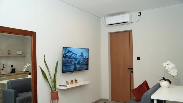 A modern living room with a flat-screen TV mounted on the wall, a potted plant, a sofa, a small table, and a wooden door. - Studio 27 Sanur - Self Check-in (Denpasar)