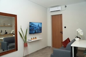 A modern living room with a flat-screen TV mounted on the wall, a potted plant, a sofa, a small table, and a wooden door. - Studio 27 Sanur - Self Check-in (Denpasar)