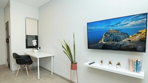 A modern room with a white desk, a potted plant, a mounted TV, and a shelf with books and figurines. - Studio 27 Sanur - Self Check-in (Denpasar)