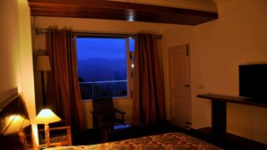 A hotel room with a bed, a chair, a desk with a television, and a window with curtains. - Vatsalyam Homestay (Shimla)