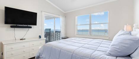 A bedroom with a bed, a TV, a ceiling fan, a window with blinds, and a view of the ocean.