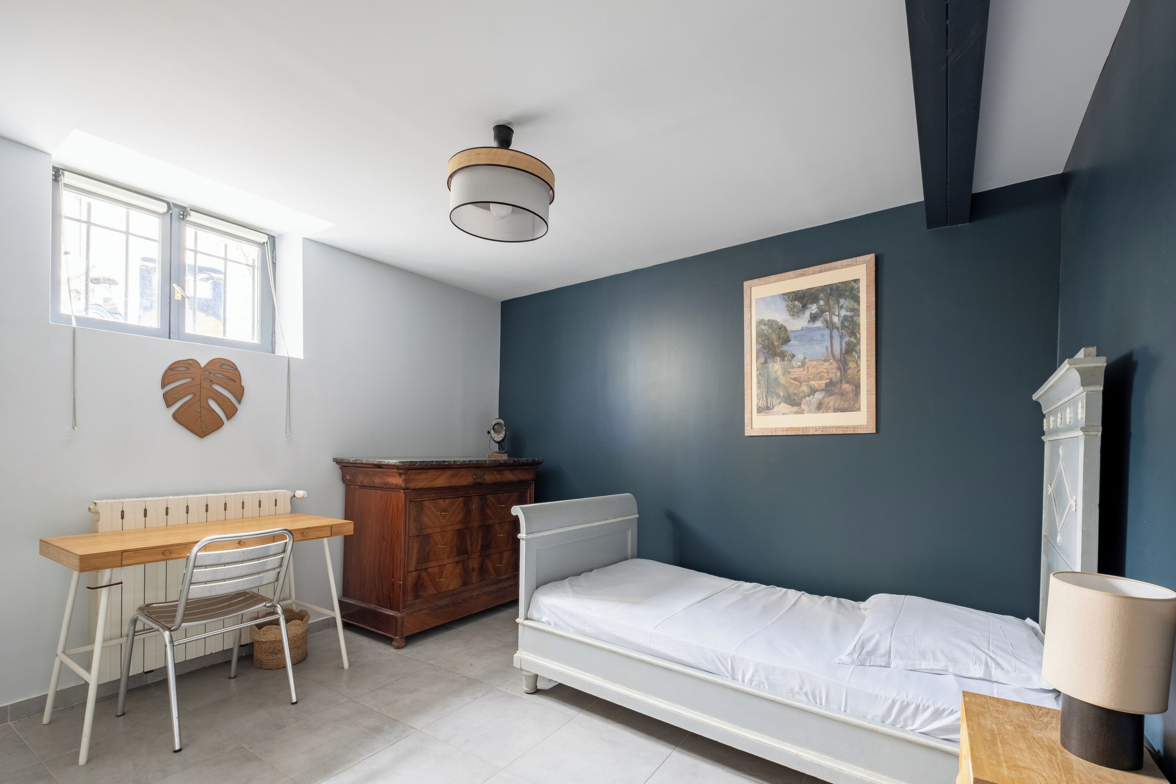 Ein Schlafzimmer mit Bett, Schreibtisch, Stuhl, Kommode, Heizkörper, Deckenleuchte und einem gerahmten Bild an der Wand.