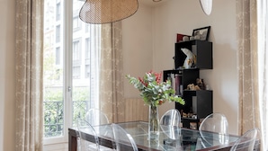 Ein Essbereich mit einem Glastisch und Stühlen, einer Vase mit Blumen sowie einem Bücherregal mit Büchern und Dekoration.