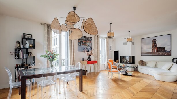 Ein modernes Wohnzimmer mit Holzdielen im Fischgrätmuster, einem Glastisch mit Stühlen, einem weißen Sofa und einem großen Gemälde an der Wand.