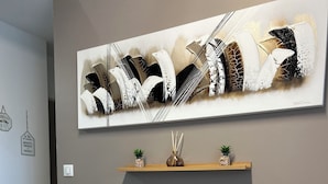 A modern living room with a grey leather sofa, two decorative pillows, a fluffy white throw, and a wall-mounted abstract art piece above a wooden shelf with small plants. - Le Cocon de la Garrigue (Agde)