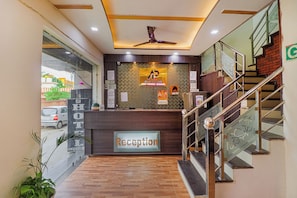 A hotel reception area with a reception desk, a ceiling fan, a staircase, and a view of the street outside. - Hotel Amishi (Haridwar)