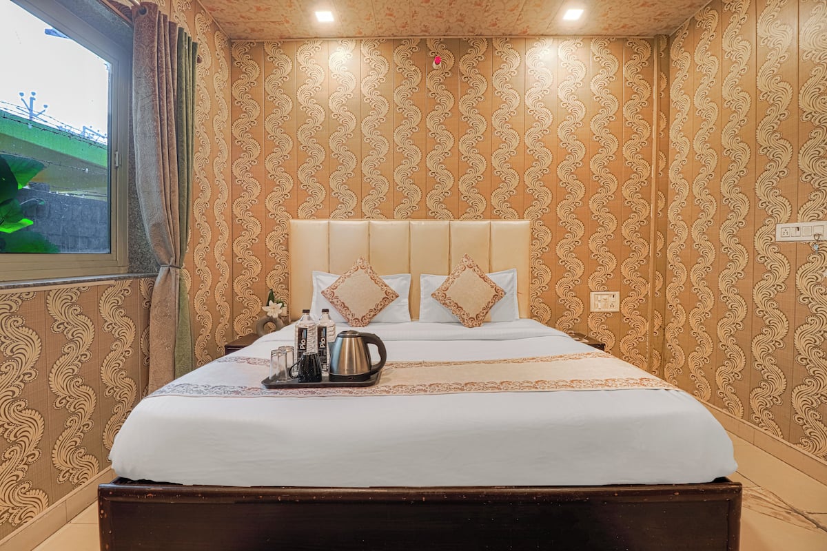 A hotel room with a large bed, a window with curtains, a wall with a patterned wallpaper, and a bedside table with a kettle and bottles.