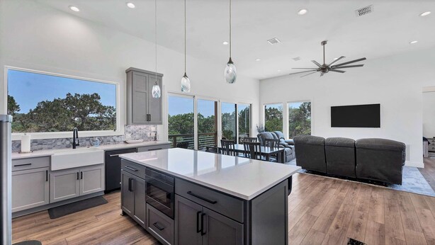 A modern kitchen with a large island, grey cabinets, and a dining area with a black leather couch and a flat-screen TV.