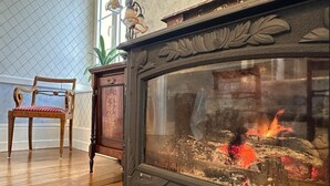 Una habitación con una silla de madera, una chimenea decorativa con puerta de cristal y un armario de madera.