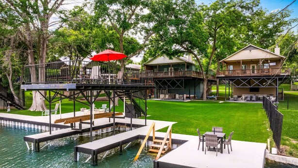 A waterfront property with a dock, outdoor seating, and a large house with a deck. - Enjoy the Full River | Fire Pit & Deck | Sleeps 12 (Seguin)