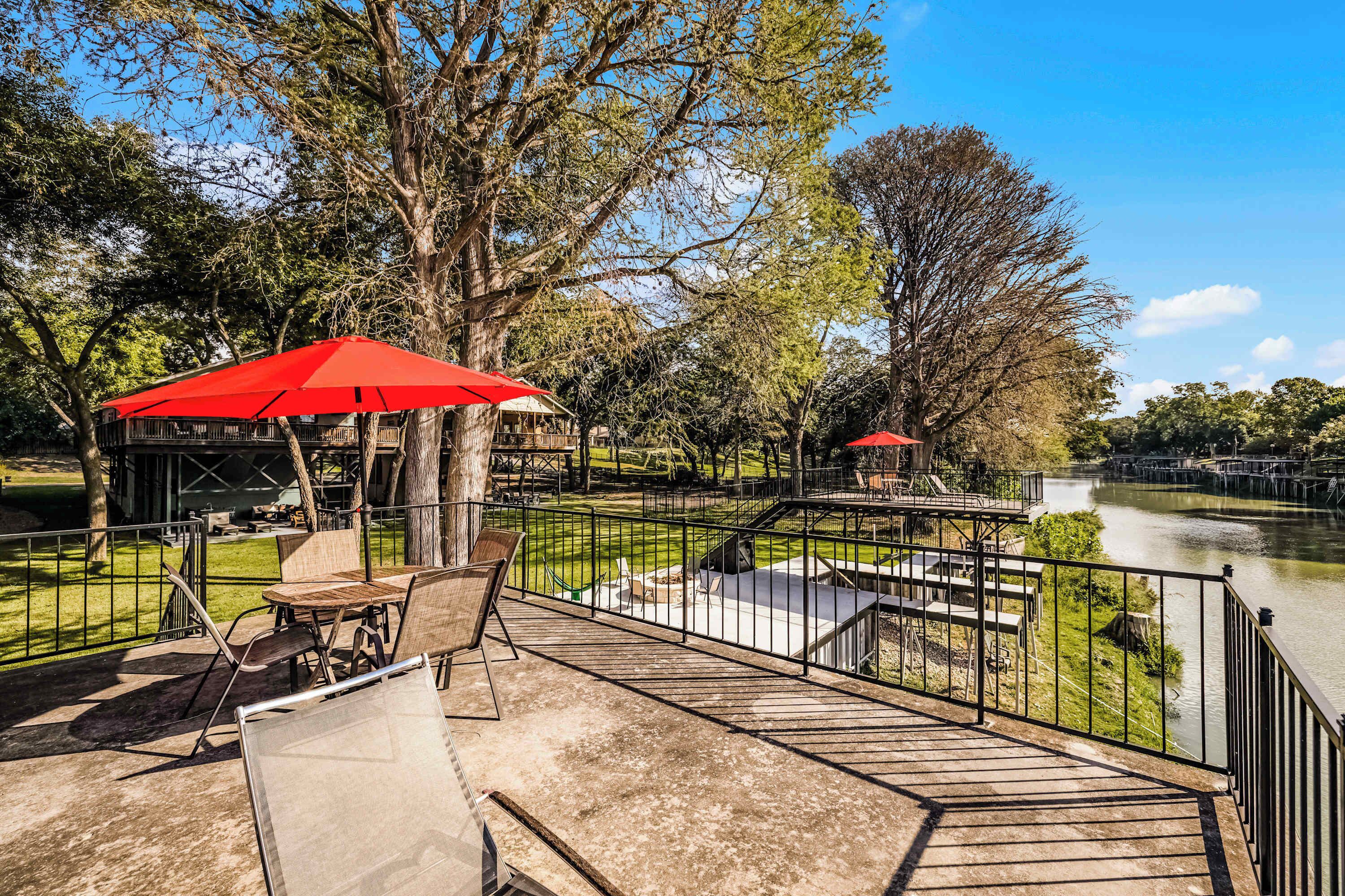 Terraza a la orilla del río con mesas al aire libre y sombrillas rojas, con vistas a un río tranquilo y abundante vegetación.