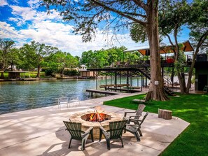 A lakeside patio with a fire pit, outdoor seating, and a dock extending into the water. - Full Lake! Waterfront Escape with Scenic Views! (Seguin)
