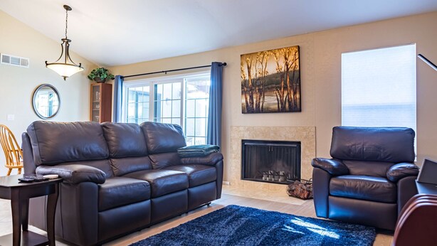 A living room with a leather sofa, a matching armchair, a fireplace, and a large window with curtains. - Cozy House, Pool, Spa, Tennis & Pickleball Courts (Tempe)