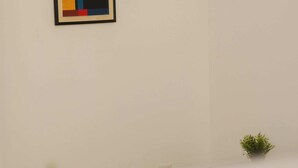 Ein Zimmer mit einem einzelnen gerahmten abstrakten Gemälde an der Wand, einer kleinen Topfpflanze und einem Fenster mit Rollläden.