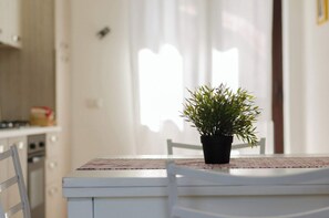 Una mesa de comedor con una planta en maceta en el centro, sillas blancas y una zona de cocina al fondo.